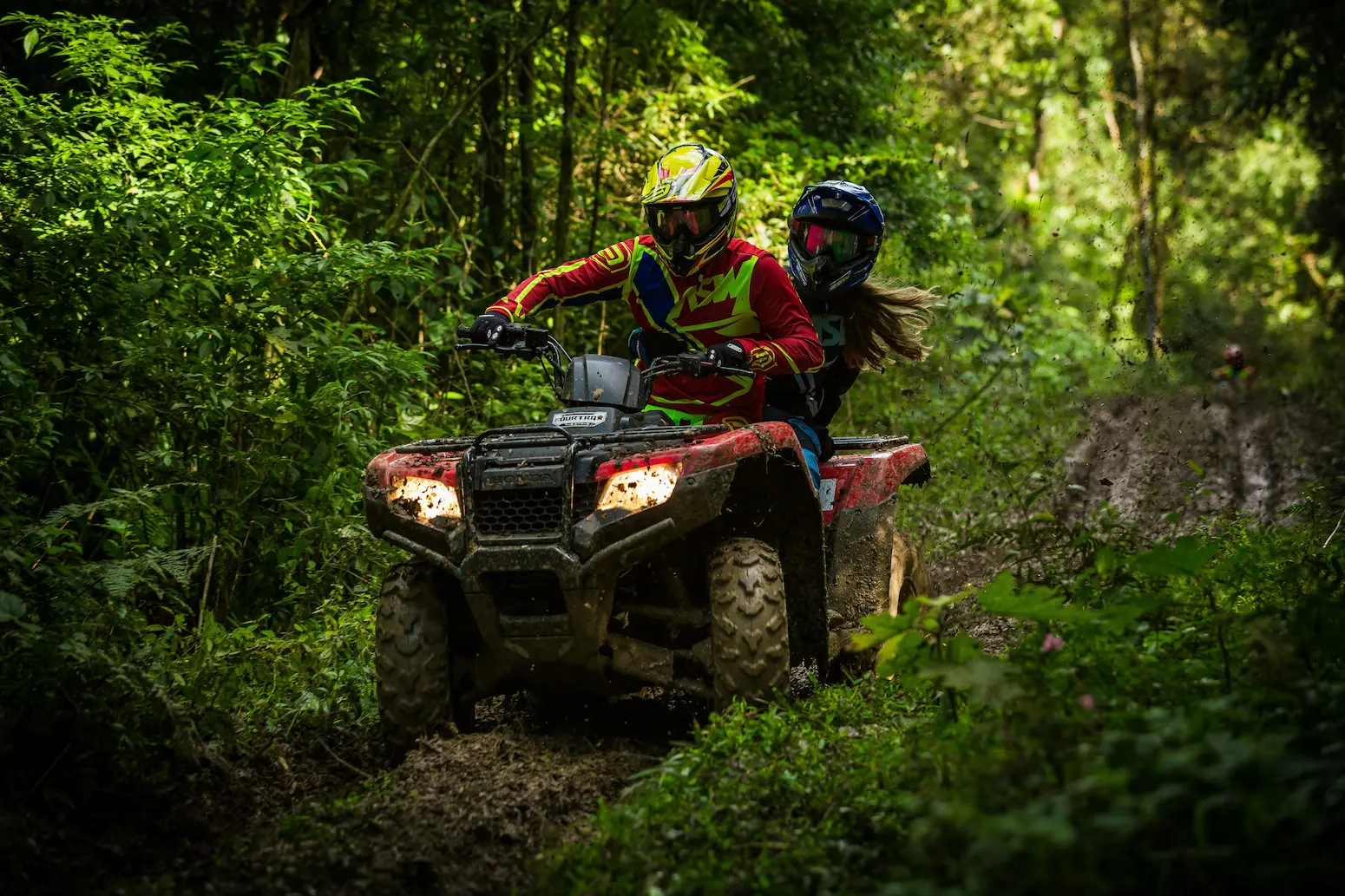 A man and woman riding a quad bike through a wooded area, surrounded by trees and grass. The vehicle is built for off-road driving, with large tires suitable for rough terrain.