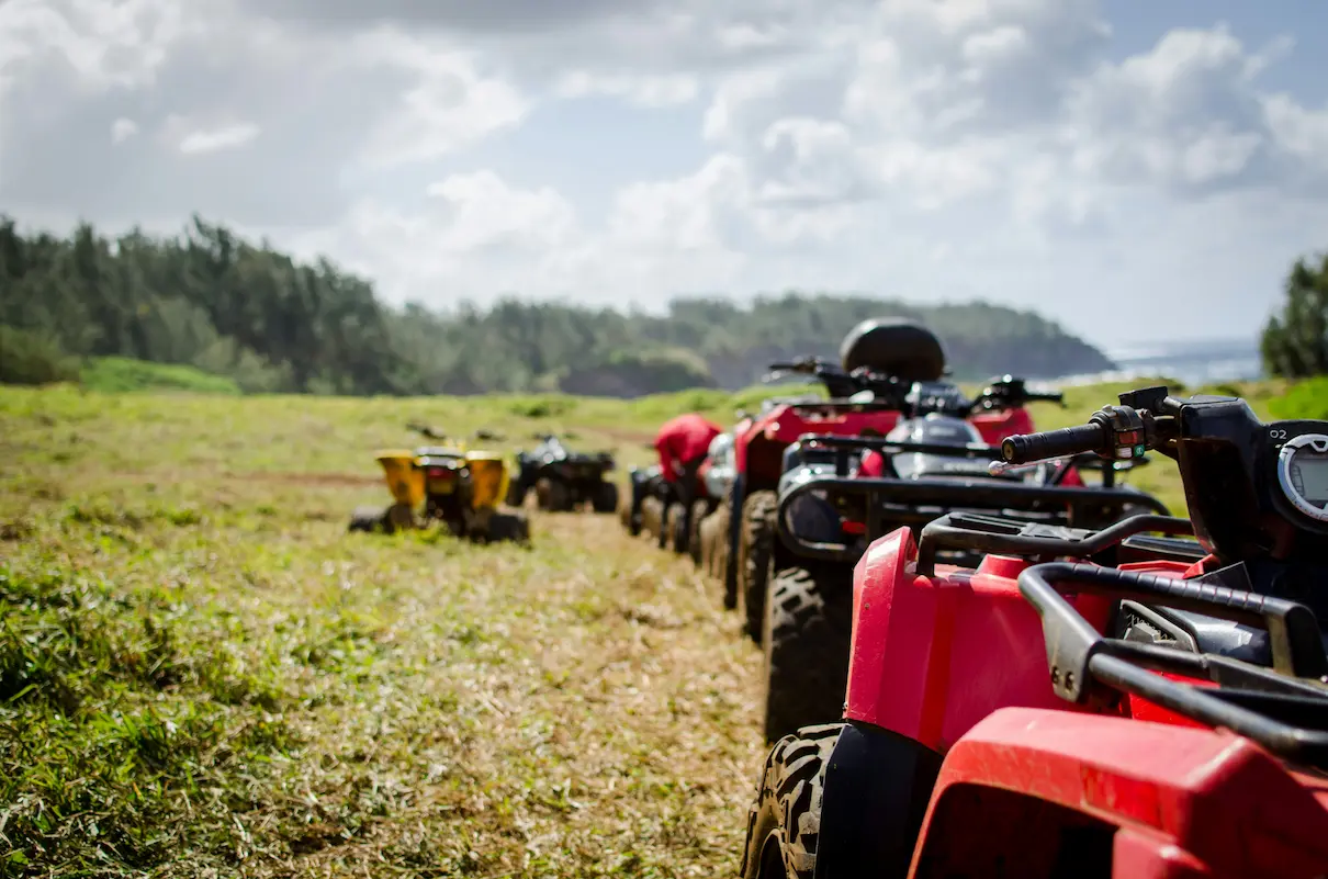 A row of quads parked in a grassy field under a partly cloudy sky, with trees in the background.