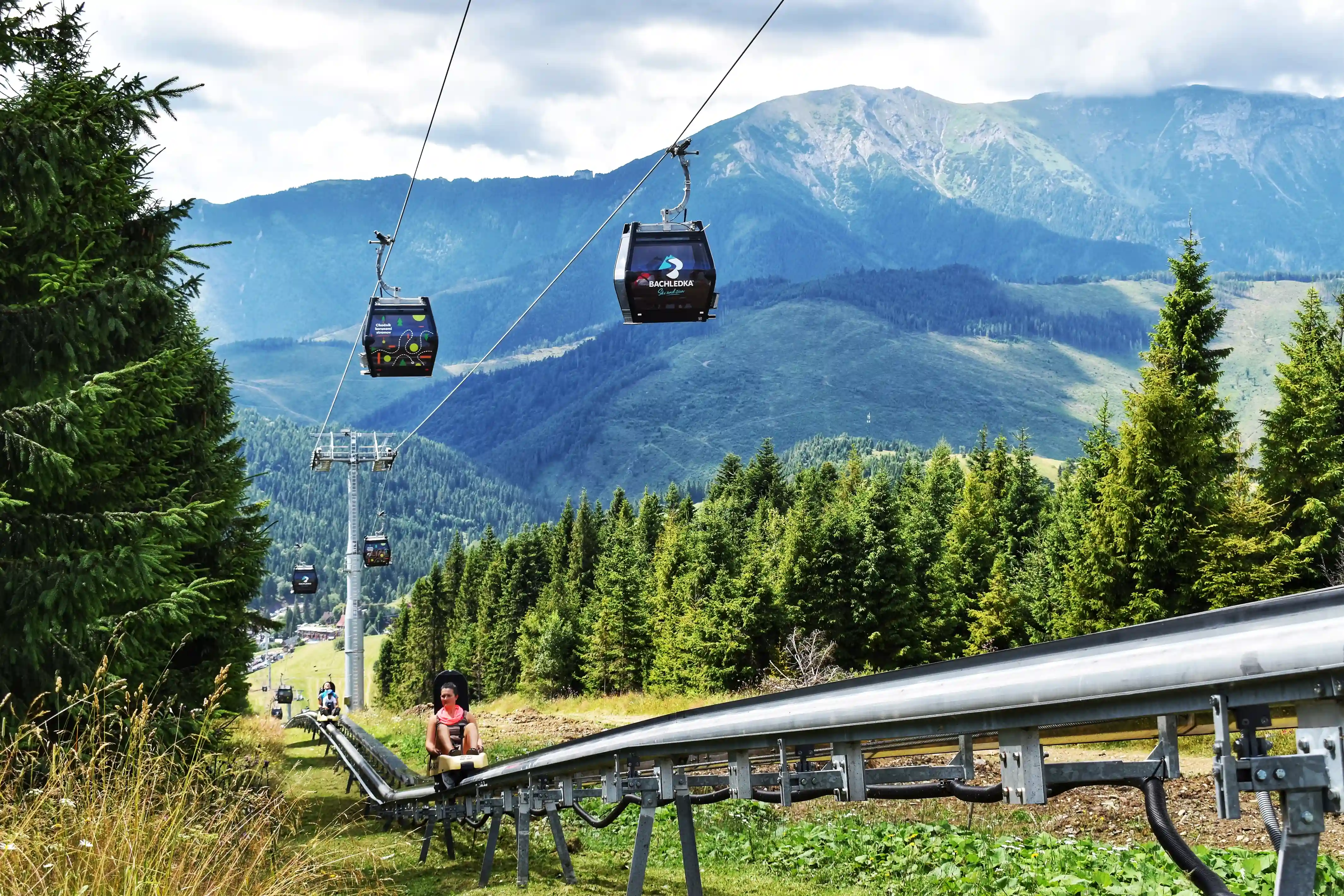 A person sitting inside a gondola, surrounded by mountains and trees, with a clear sky and clouds in the background. The gondola is part of a ski lift at a mountain resort.