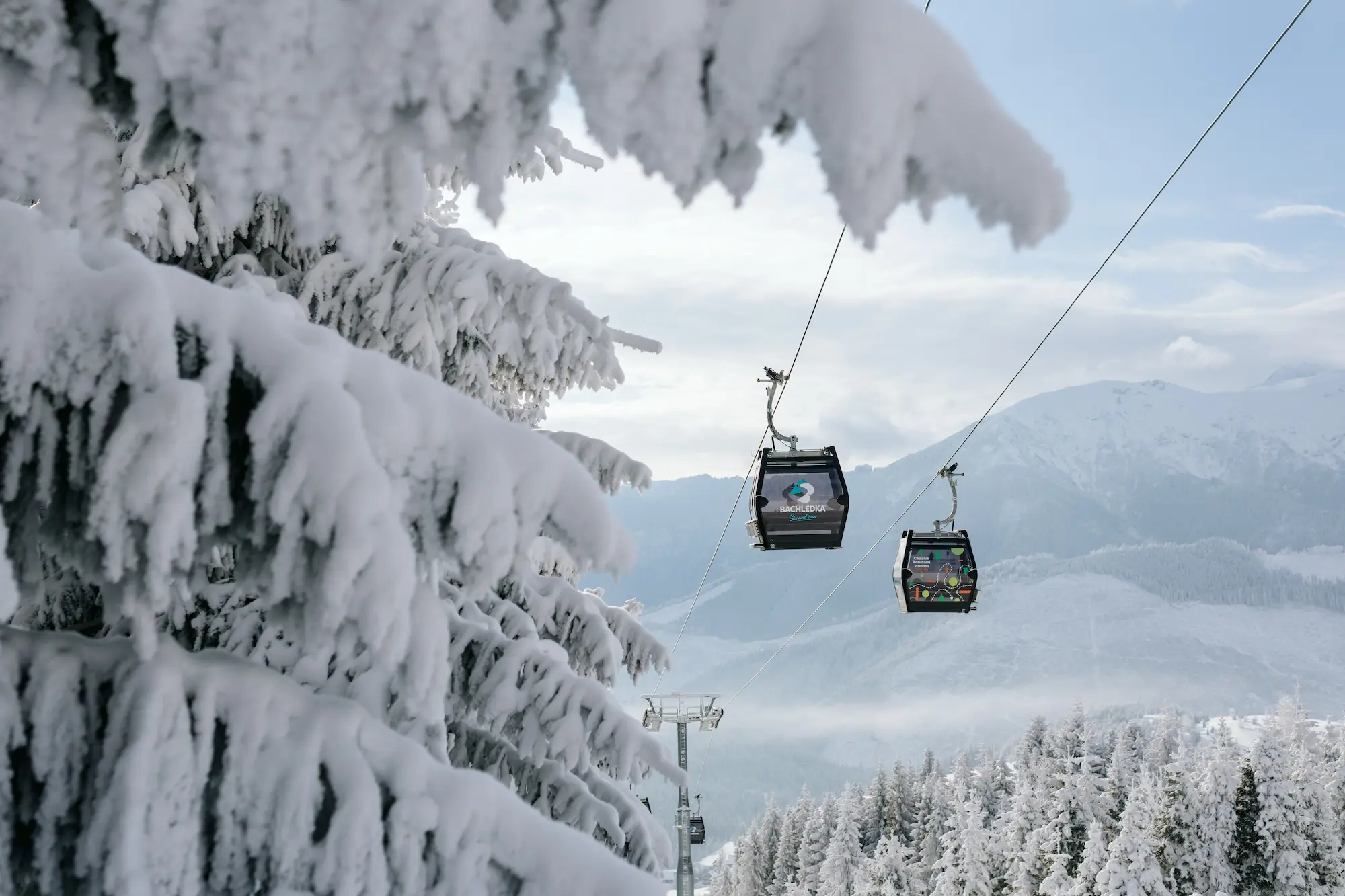 Osoba siedząca w gondoli, otoczona górami i drzewami, z czystym niebem i chmurami w tle. Gondola jest częścią wyciągu narciarskiego w stacji górskiej.