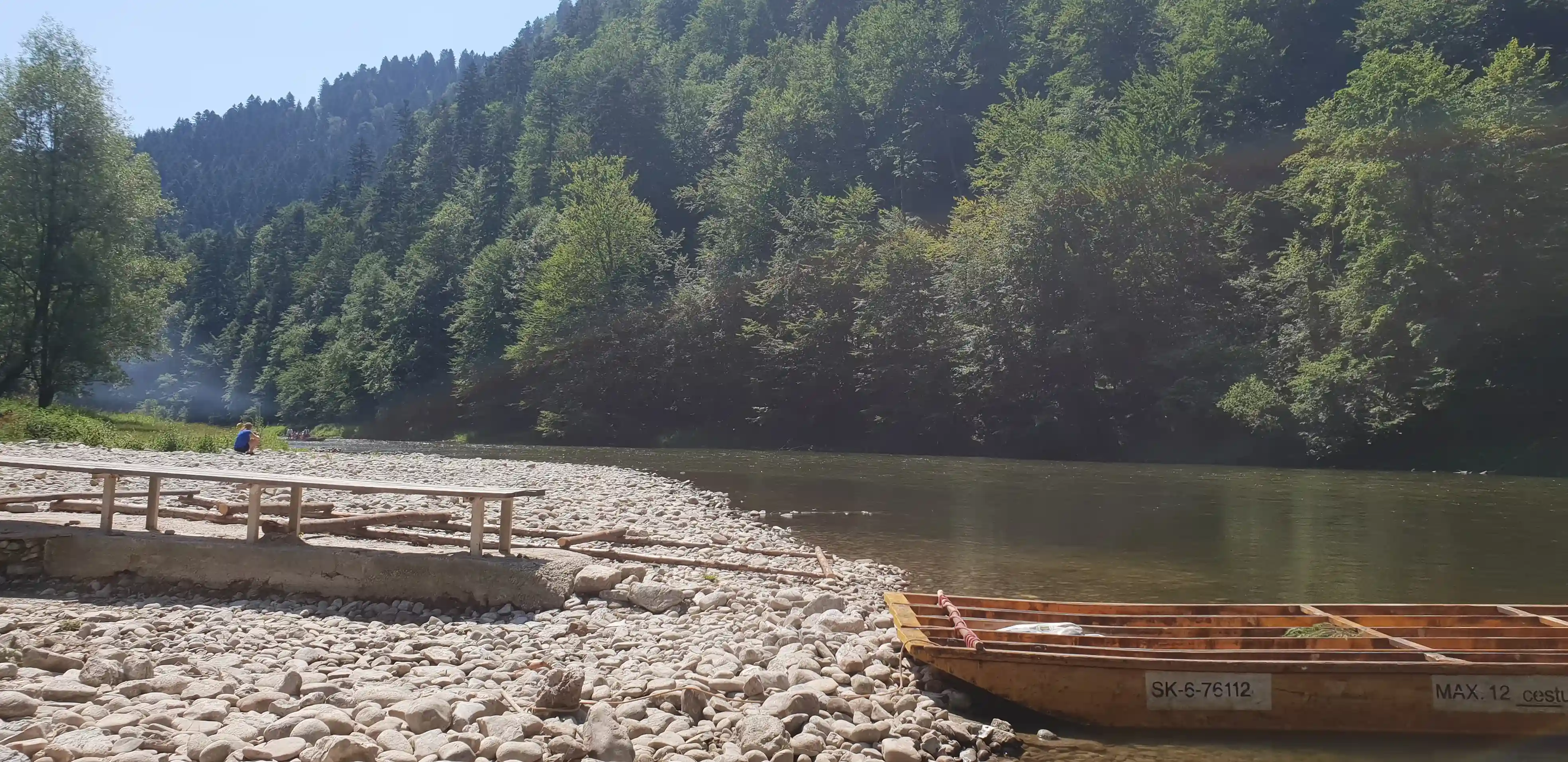 A boat resting on a rocky shore beside a river, surrounded by trees and mountains under a clear sky.