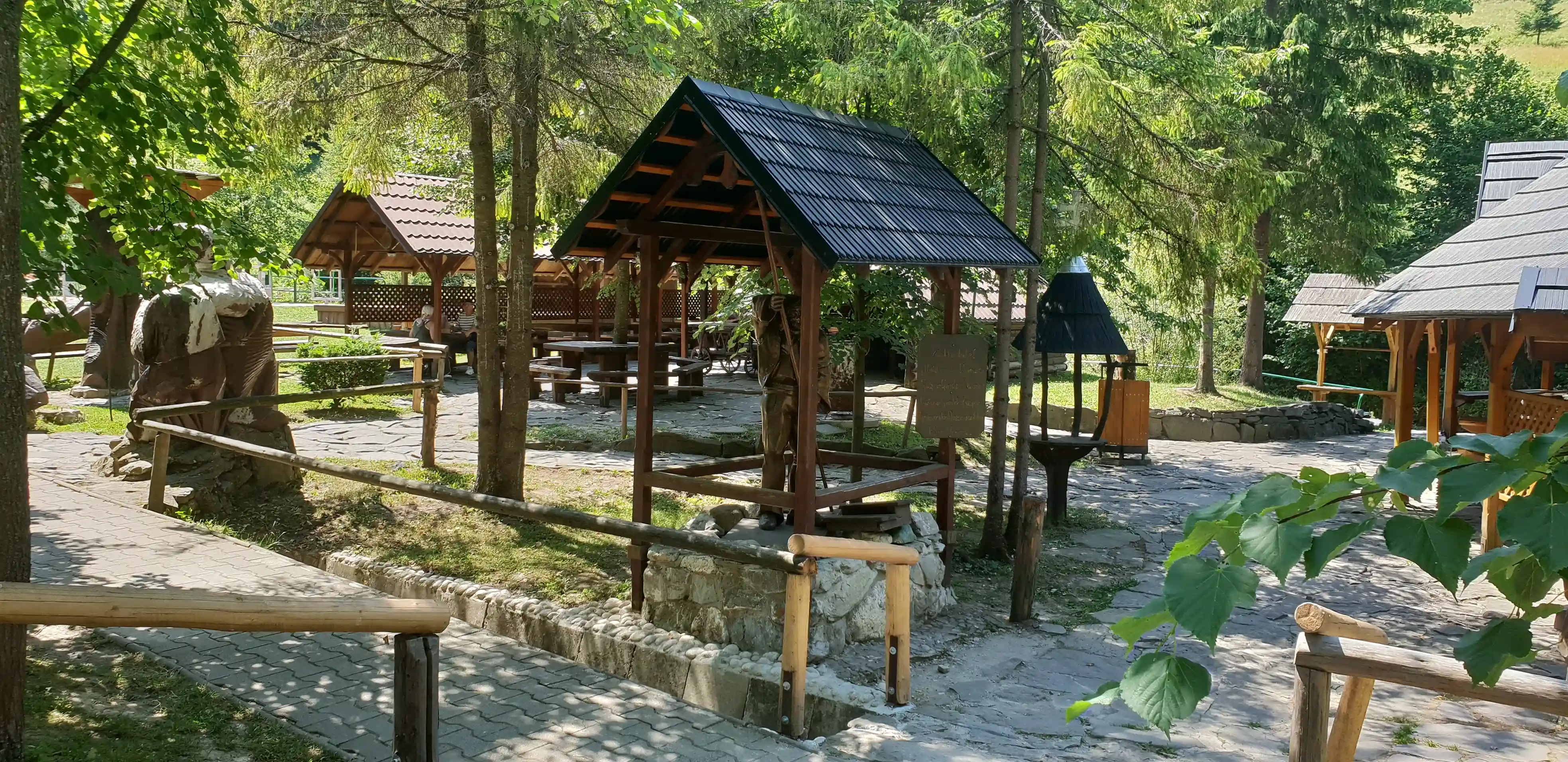 A scenic park featuring a gazebo surrounded by trees and plants, with a grassy ground and a wooden fence nearby. The gazebo is made of wood, resembling a log cabin structure.