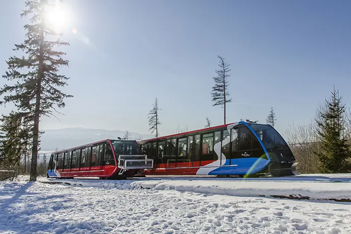 Czerwono-niebieska kolejka linowo-terenowa wjeżdżająca na Hrebienok w słoneczny, zimowy dzień na tle tatrzańskich sosen.